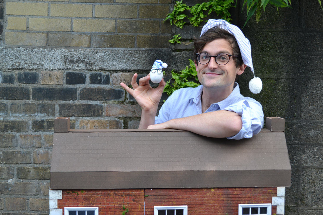 Production image from GOODNIGHT EGG. A man wearing glasses, dressed in a blue nightshirt and striped blue nightcap holds a book in one hand and a white egg in the other. The white egg is also wearing tiny glasses and a tiny blue nightcap. In the background, there are cardboard moving boxes. A wooden model house can be partially seen behind the man.