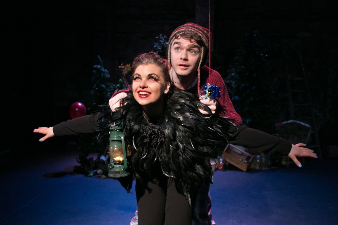 Production image from HALF LIGHT. A boy wearing a red jumper and woolly hat holds the string of a red balloon in one hand and a green lantern in the other. He is hanging on to the back of a woman costumed as a crow, who has her arms outstretched as if she they are flying through the air. The background is dark.