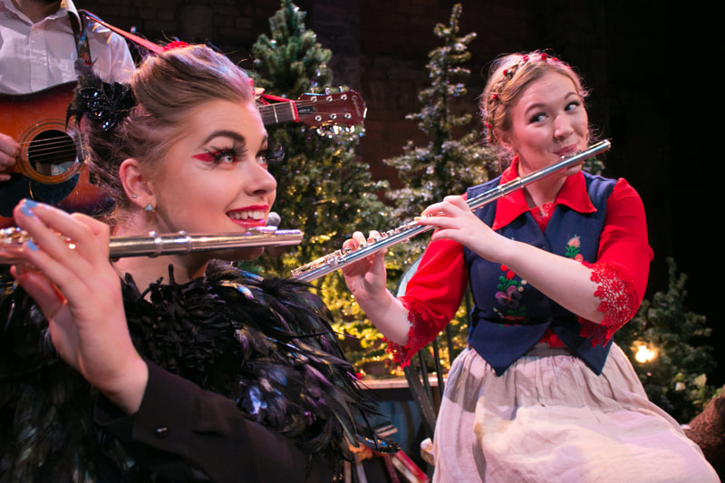 Production image from HALF LIGHT. Two women sit playing flutes. One is dressed as a crow, with a top made of black feathers, and black feathers in her hair. She is wearing thick black false eyelashes and thick pink eyeliner. The other woman is wearing a red blouse, blue waistcoat and beige skirt and has her hair in plaits. In the background, there are two faux Christmas trees lit from behind with amber light.